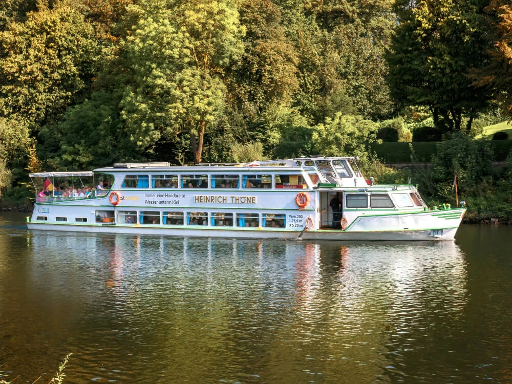 Weiße Flotte, Mülheim an der Ruhr Das Schiff "Heinrich Thöne" der Weißen Flotte fährt auf einem ruhigen Fluss durch grünen Wald.