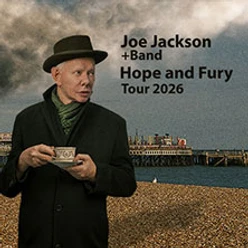 Musiker Joe Jackson mit Hut hält eine Tasse, im Hintergrund ein Pier.