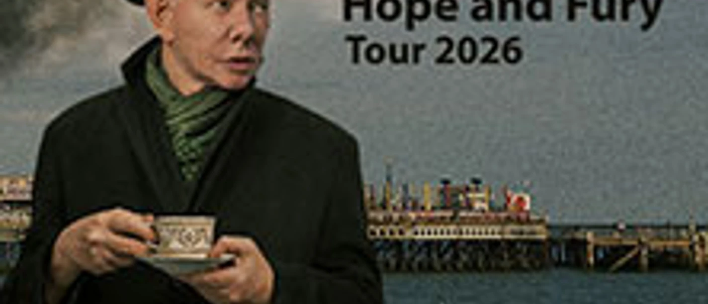 Musiker Joe Jackson mit Hut hält eine Tasse, im Hintergrund ein Pier.