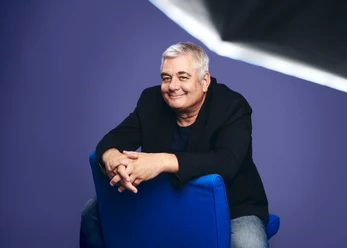 Pressefoto 3 Ingo Oschmann HerzScherz Fotograf Jean Ferry.jpg Mann mit grauen Haaren sitzt lächelnd auf blauem Stuhl vor violettem Hintergrund im Studio.
