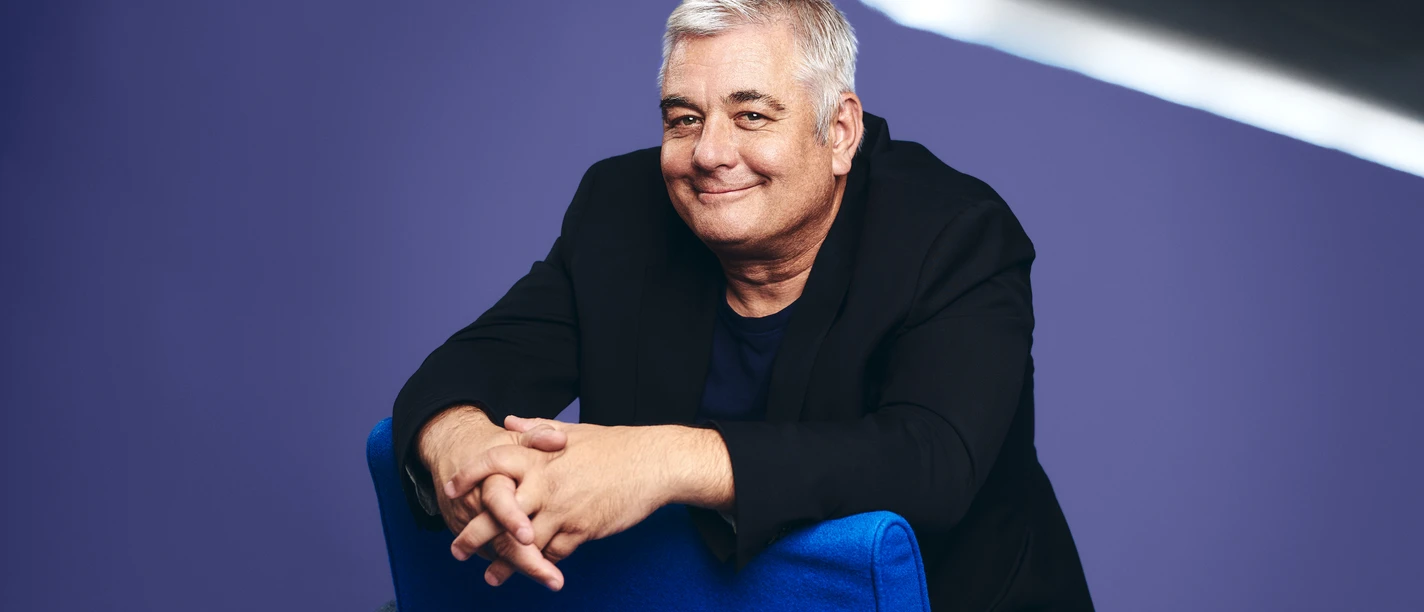 Pressefoto 3 Ingo Oschmann HerzScherz Fotograf Jean Ferry.jpg Mann mit grauen Haaren sitzt lächelnd auf blauem Stuhl vor violettem Hintergrund im Studio.
