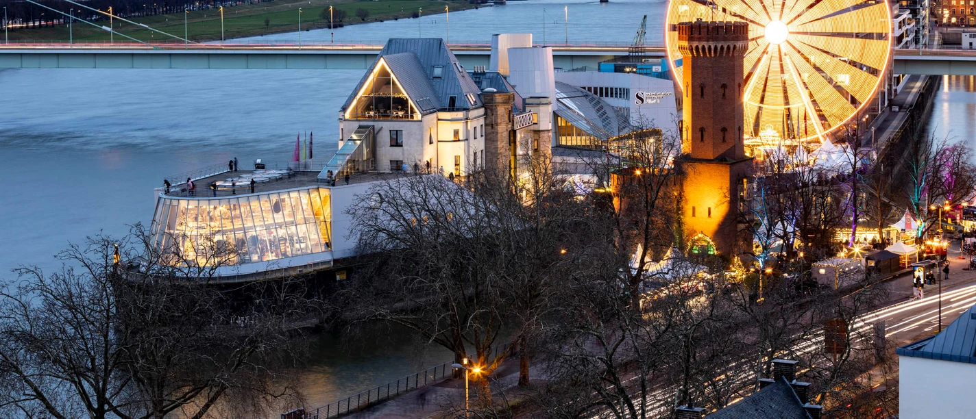View of the brightly lit Christmas market at the Cologne Chocolate Museum, with the Rhine in the background.