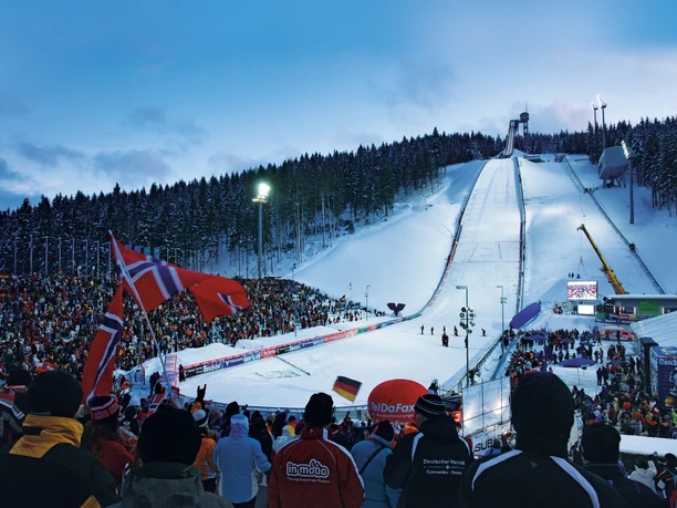 3-sparkasse-vogtland-arena-zur-freien-verwendung.jpg Skisprung-Arena bei Abenddämmerung, umgeben von schneebedecktem Wald, mit jubelnden Zuschauern.
