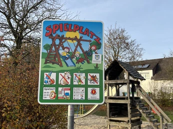 Spielplatz am Schneiderberg Hinweisschild mit Spielplatzregeln vor Holz-Klettergerüst und Rutsche im sandigen Spielbereich.