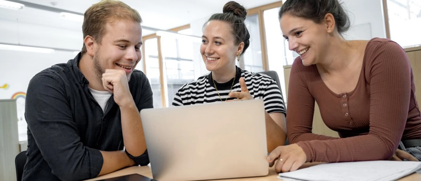 Bild 1.jpeg Drei Studierende sitzen an einem Tisch und arbeiten gemeinsam an einem Laptop in heller Umgebung.