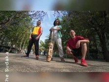 Drei Personen posieren draußen bei einem Spaziergang auf einem baumbestandenen Platz.