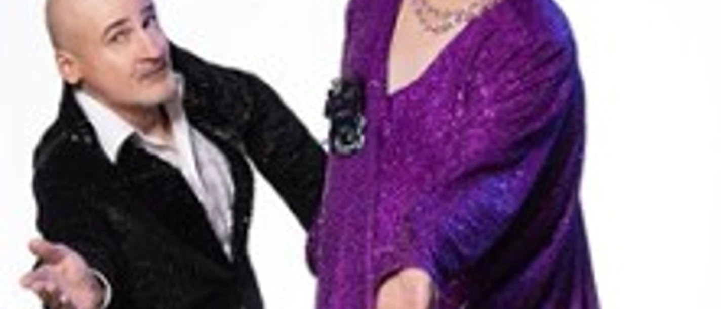 Zwei Personen in glamourösen Outfits lächeln und posieren auf einem hellen Hintergrund.