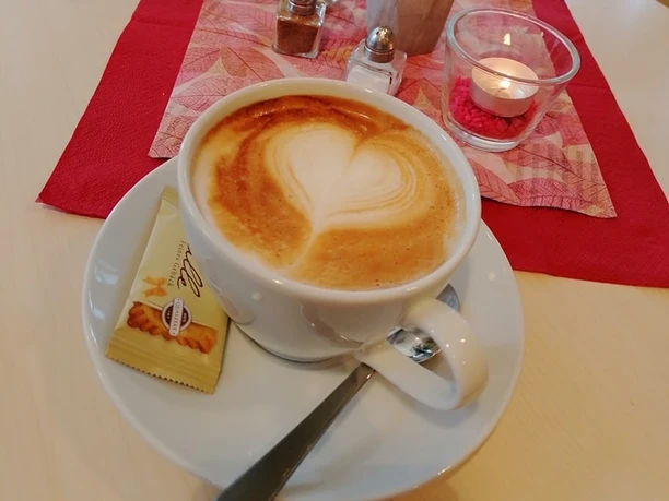 Kaffee.jpg Ein Cappuccino mit herzförmiger Latte Art auf einem Tisch mit roten Servietten, Blumen und Teelicht.