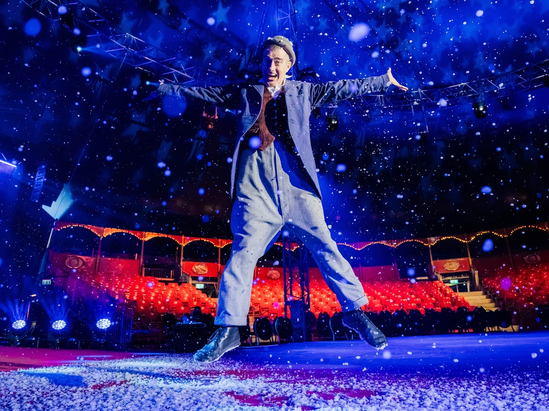 Cologne Christmas Circus Ein Akrobat im Zirkuszelt springt freudig in die Luft, umgeben von fallendem Kunstschnee.An acrobat in the circus tent leaps joyfully into the air, surrounded by falling artificial snow.