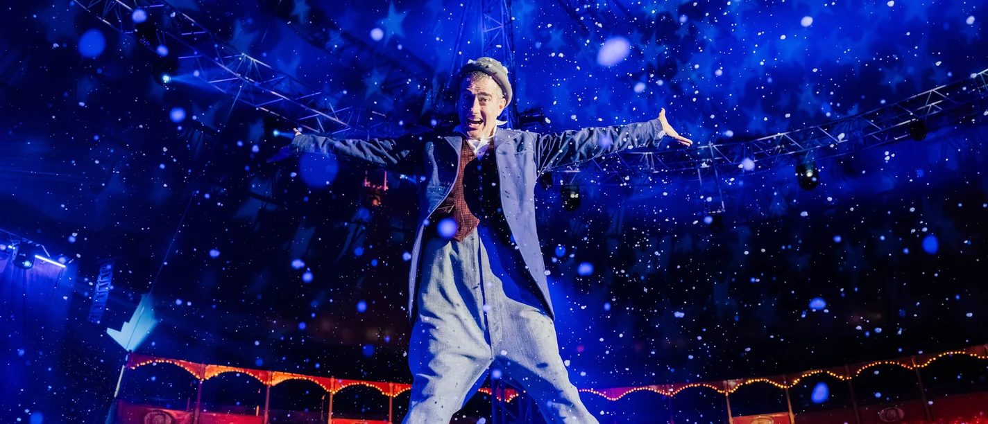 Koelner-WeihnachtsCircus Ein Akrobat im Zirkuszelt springt freudig in die Luft, umgeben von fallendem Kunstschnee.