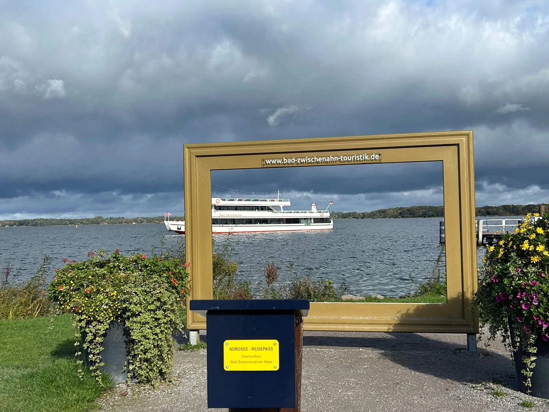 Stempelstelle-Nordsee-Reisepass-Goldener-Bilderrahmen-Bad-Zwischenahn.jpg Großer goldener Bilderrahmen mit Blick auf das Zwischenahner Meer und ein vorbeifahrendes Schiff.