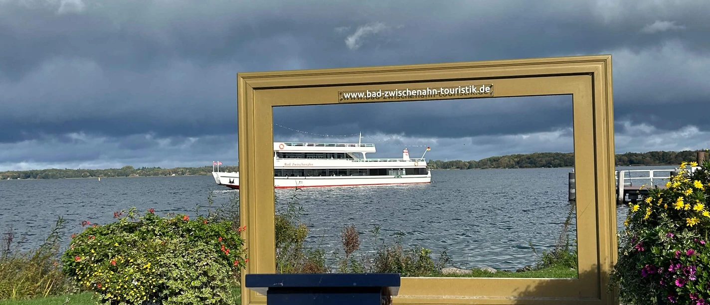 Stempelstelle-Nordsee-Reisepass-Goldener-Bilderrahmen-Bad-Zwischenahn.jpg Großer goldener Bilderrahmen mit Blick auf das Zwischenahner Meer und ein vorbeifahrendes Schiff.