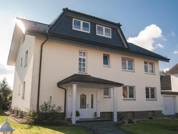 Außenansicht 2 Zweistöckiges Wohnhaus mit hellem Putz, dunkelgrauem Dach und überdachtem Eingang mit Säulen.
