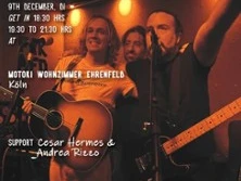 Drei Musiker auf Bühne mit Gitarren und Mikrofonen, in warmem Licht bei einem Live-Auftritt.