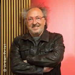Ein lächelnder älterer Mann mit Brille und lockigem Haar, trägt eine Lederjacke.
