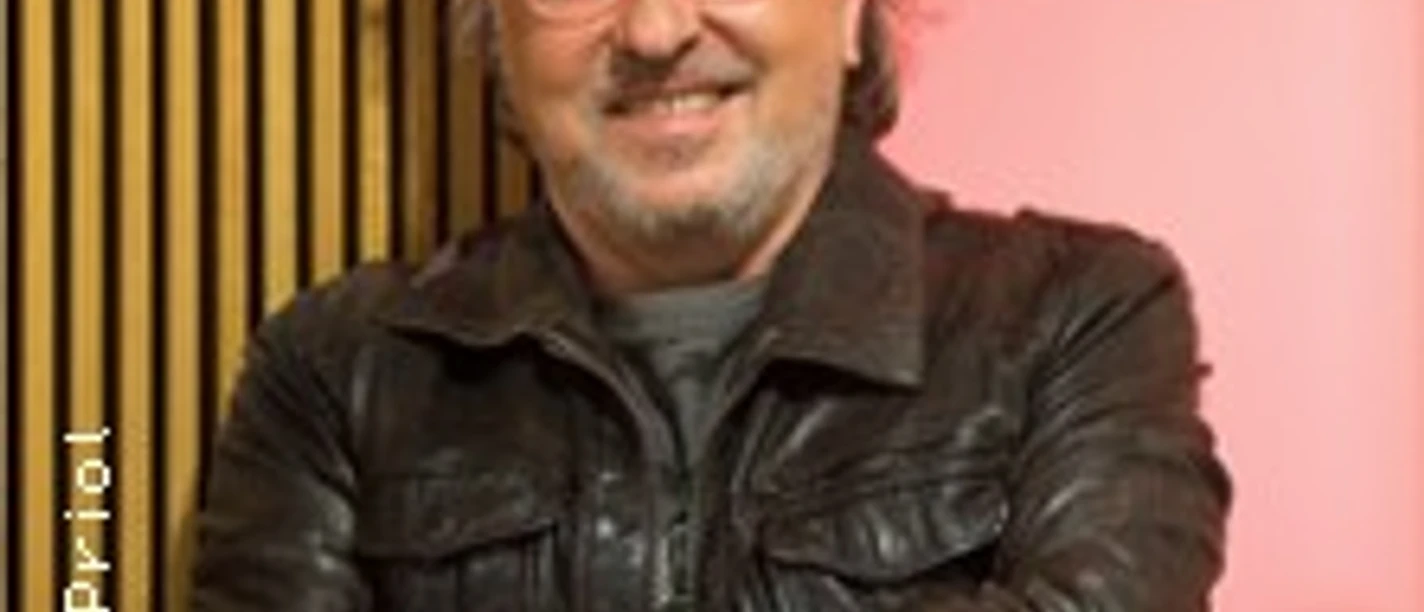 Ein lächelnder älterer Mann mit Brille und lockigem Haar, trägt eine Lederjacke.