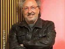 Ein lächelnder älterer Mann mit Brille und lockigem Haar, trägt eine Lederjacke.