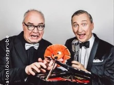 Zwei Männer im Anzug mit Mikrofon und Requisiten posieren humorvoll.