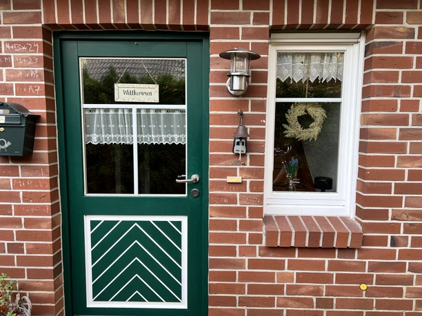 Hauseingang(1).jpg Grüner Hauseingang mit Glastür, rotem Klinkermauerwerk, Fenster, Blumenkranz und Willkommensschild.