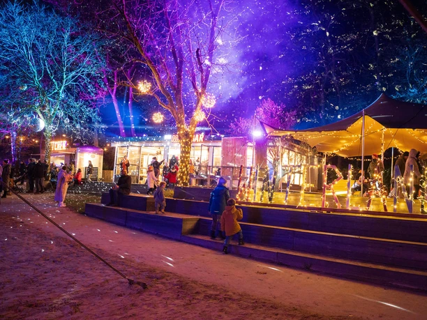 Blackfoot Winterland Ein festlich erleuchteter Wintermarkt mit farbigen Lichtern, umgeben von schneebedeckten Bäumen.