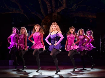 Riverdance 30 – Die neue Generation Riverdance-Tänzerinnen in präziser Pose auf der Bühne, umhüllt von kräftigem Licht in lebhaften Farben.Riverdance dancers in precise poses on stage, enveloped by powerful light in vivid colors.