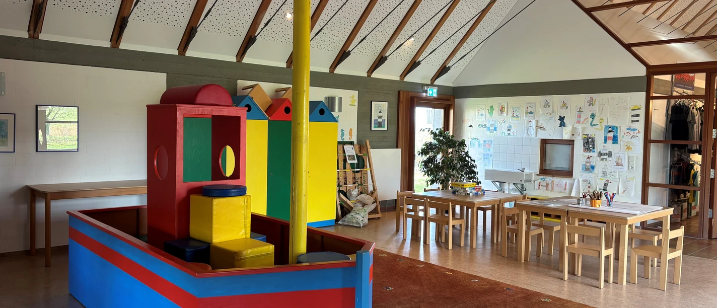 Kinderspielzimmer Wremen Helles Spielzimmer mit buntem Holzschiff, Maltischen und Kindersitzecke unter hohem Dach.