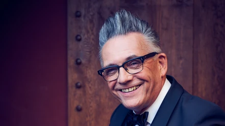 Götz Alsmann mit Brille und Fliege lächelt vor einer Holzwand.