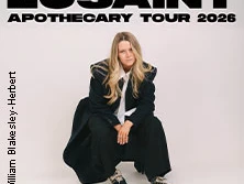 Frau mit langem Haar in dunklem Mantel und Sneakers vor weißem Hintergrund, Luisaint Tour 2026.