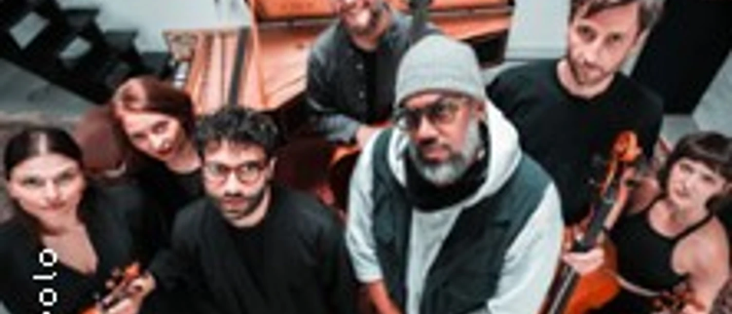 Eine Musikgruppe steht in einem Studio mit Instrumenten wie Geige und Kontrabass.