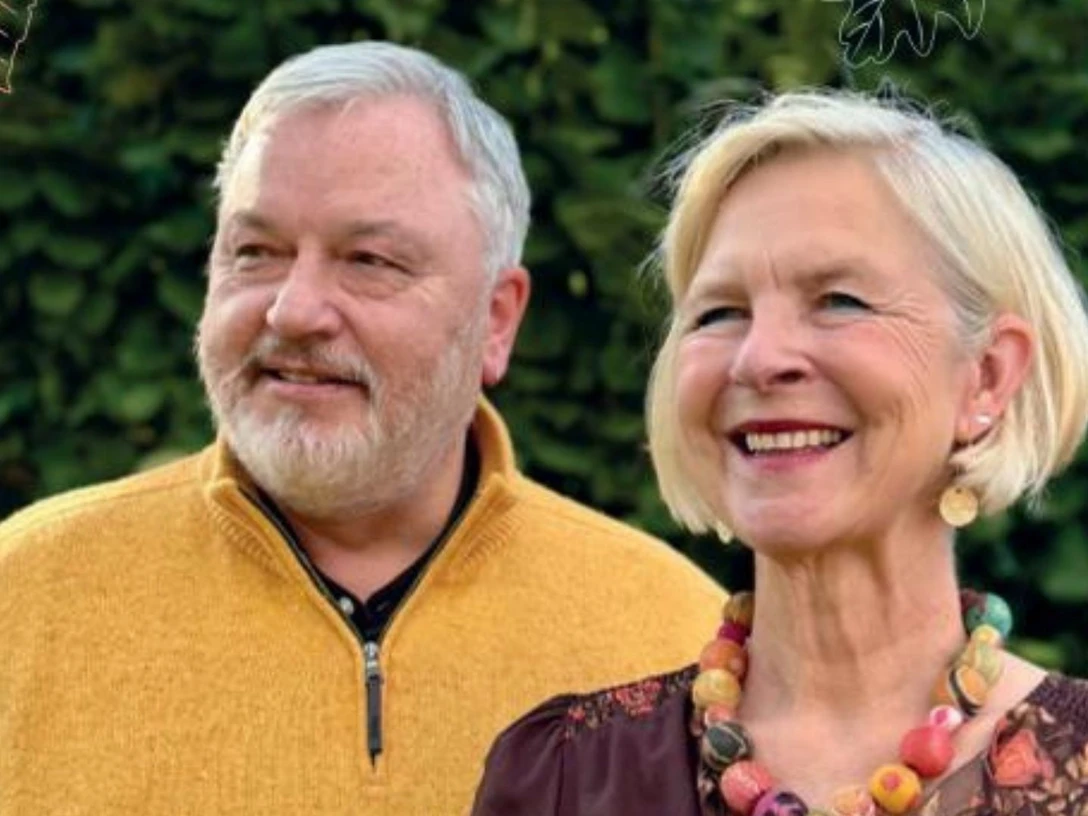 Finissage Event-Bild.jpg Du siehst zwei Menschen draußen. Sie lächeln. Hinter ihnen ist grüne Hecke.