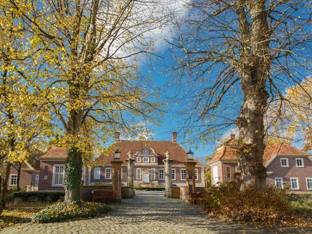 Schloss Herzford Backsteinernes Schloss mit rotem Walmdach, flankiert von Bäumen mit Herbstlaub unter blauem Himmel.