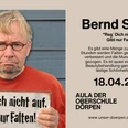 Bernd Stelter.png Ein Mann mit rotem Pullover und rundem Brillengestell hält ein Schild mit humorvollem Spruch.