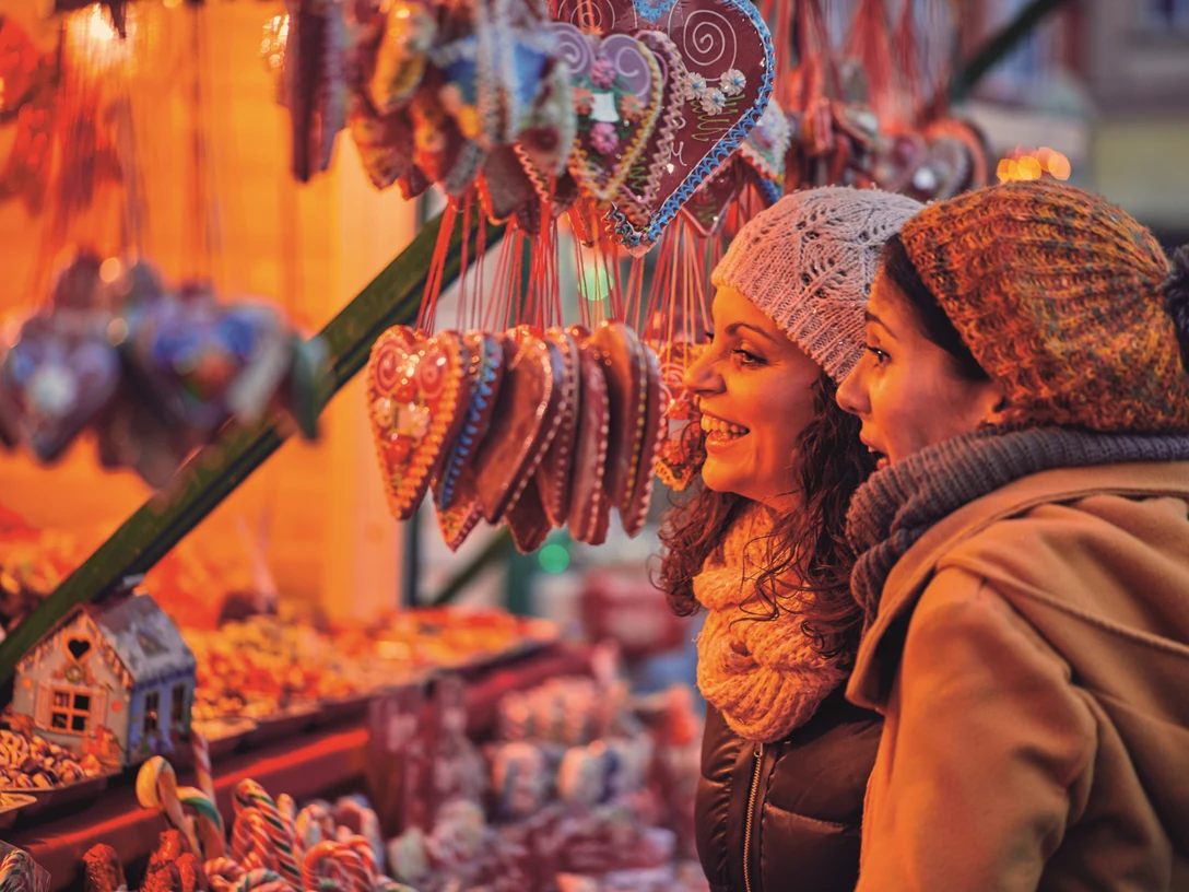 Otterndorfer Sternenmarkt Zwei Frauen betrachten lächelnd Lebkuchenherzen an einem festlich beleuchteten Weihnachtsmarktstand.