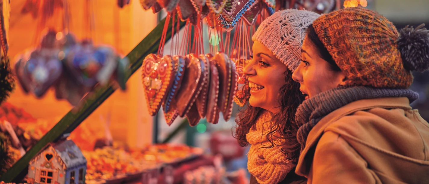 Otterndorfer Sternenmarkt Zwei Frauen betrachten lächelnd Lebkuchenherzen an einem festlich beleuchteten Weihnachtsmarktstand.