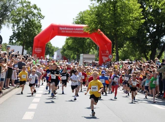 Stadtlauf_3626-os A 1366.jpg Kinder starten beim Stadtlauf unter rotem Torbogen, umgeben von jubelnden Zuschauerreihen.