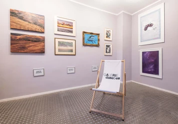 Galerie_daneben Ein kleiner Raum mit Kunstwerken an den Wänden und einem weißen Klappstuhl mit dem Schriftzug "Galerie daneben".