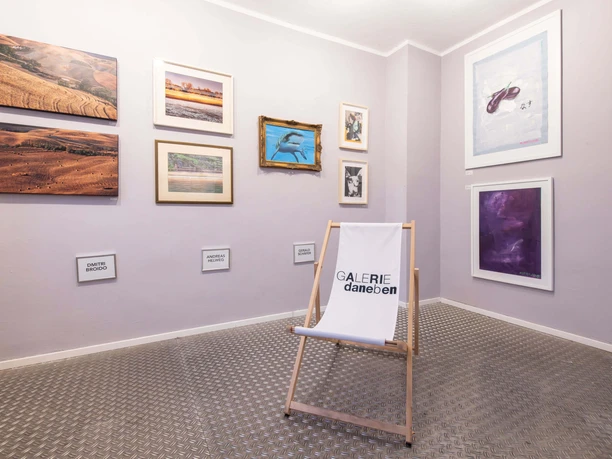 Galerie_daneben Ein kleiner Raum mit Kunstwerken an den Wänden und einem weißen Klappstuhl mit dem Schriftzug "Galerie daneben".