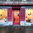 Galerie_daneben Galerieeingang mit rotem Holztor zwischen zwei großen Schaufenstern mit Kunstwerken.Gallery entrance with red wooden door between two large display windows with works of art.