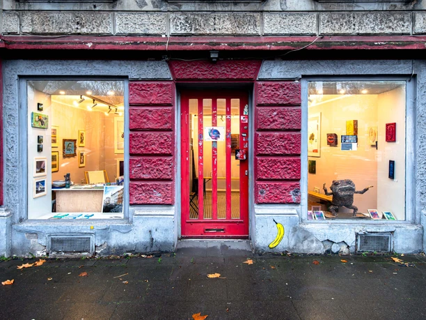 Galerie_daneben Galerieeingang mit rotem Holztor zwischen zwei großen Schaufenstern mit Kunstwerken.