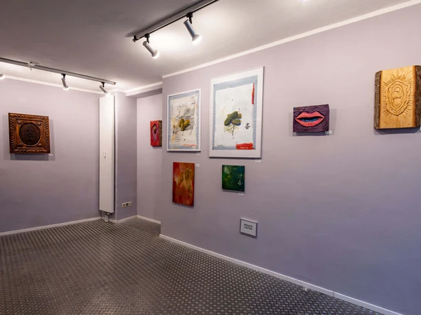 Galerie_daneben In einem kunstvoll beleuchteten Raum hängen diverse Gemälde und Skulpturen auf hell-lila Wänden.