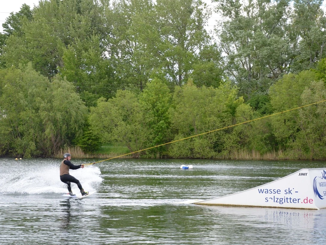 Wasserskianlage_Salzgittersee (4)_HD_16zu9.JPG Person fährt Wakeboard am Seilzug über den Salzgittersee vor grünem Ufer mit Bäumen.