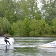 Wasserskianlage_Salzgittersee (4)_HD_16zu9.JPG Person fährt Wakeboard am Seilzug über den Salzgittersee vor grünem Ufer mit Bäumen.