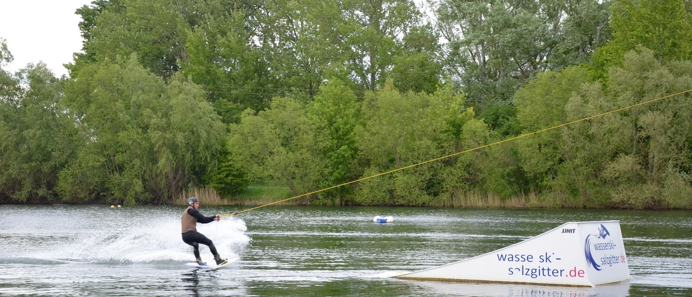 Person fährt Wakeboard am Seilzug über den Salzgittersee vor grünem Ufer mit Bäumen.