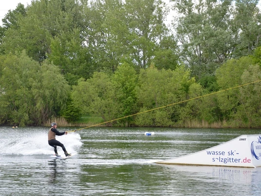 Wasserskianlage_Salzgittersee (4)_HD_16zu9.JPG Person fährt Wakeboard am Seilzug über den Salzgittersee vor grünem Ufer mit Bäumen.