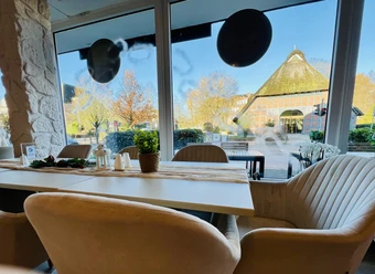 Blick aus modernem Café mit hellen Sesseln auf Fachwerkhaus mit Reetdach und herbstlichen Bäumen.