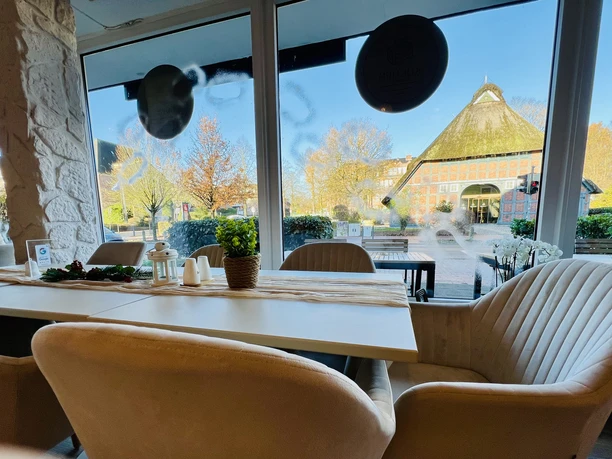 Im Café Blick aus modernem Café mit hellen Sesseln auf Fachwerkhaus mit Reetdach und herbstlichen Bäumen.