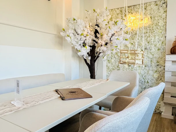 Sitzbereich im Café Heller Cafébereich mit weißen Möbeln, Blütenbaum, floraler Wanddekoration und Hängeschaukel.