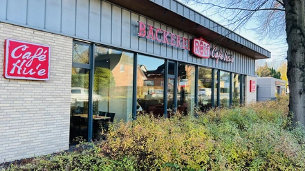 Moderne Bäckerei mit großen Glasfronten, rotem Schriftzug „Bäckerei Rolf Café Hu'e“ und Grünbepflanzung.
