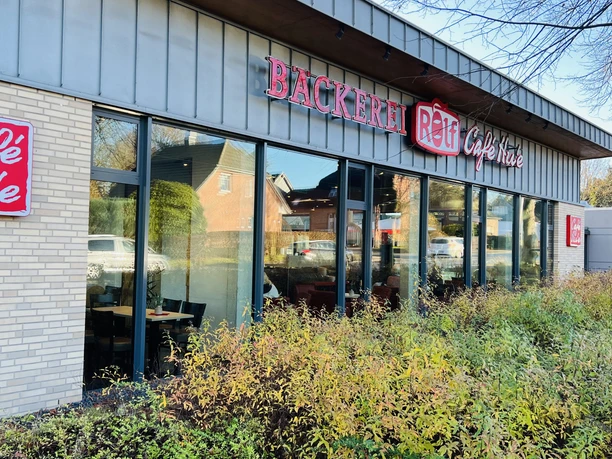 Moderne Bäckerei mit großen Glasfronten, rotem Schriftzug „Bäckerei Rolf Café Hu'e“ und Grünbepflanzung.