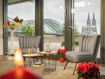 X-mas im the view Cologne Blick durch eine Fensterfront auf die Hohenzollernbrücke und den Dom Kölns, flankiert von stilvollen Sesseln und weihnachtlicher Dekoration.View through a window front onto the Hohenzollern Bridge and Cologne Cathedral, flanked by stylish armchairs and Christmas decorations.